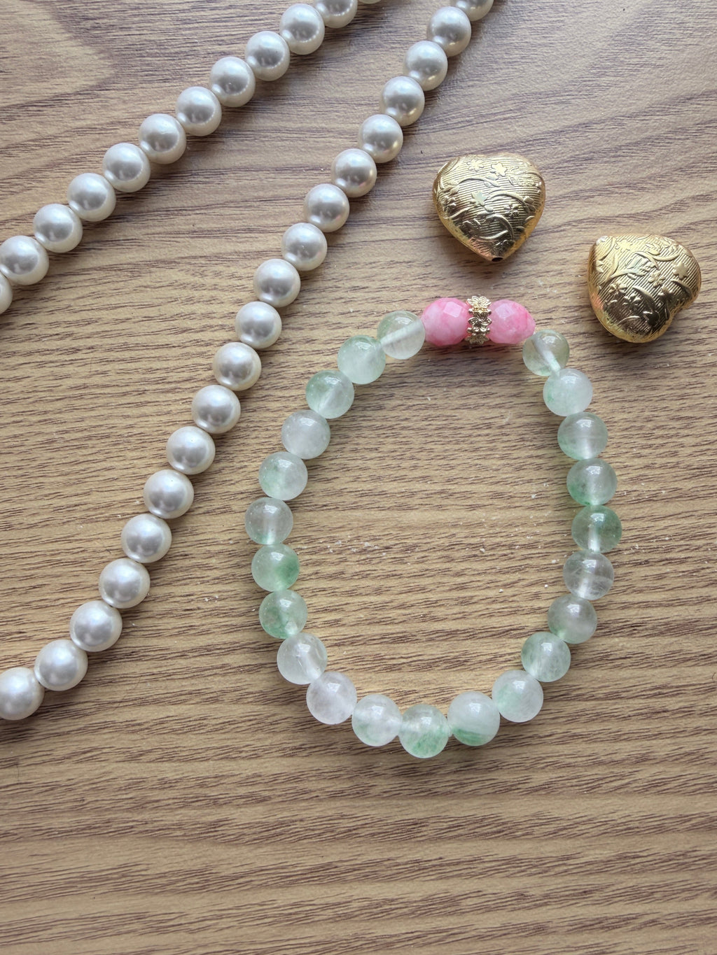"Lifestyle shot of mint green jade and candy bracelet styled with pearl necklace and gold heart accessories on wood surface"