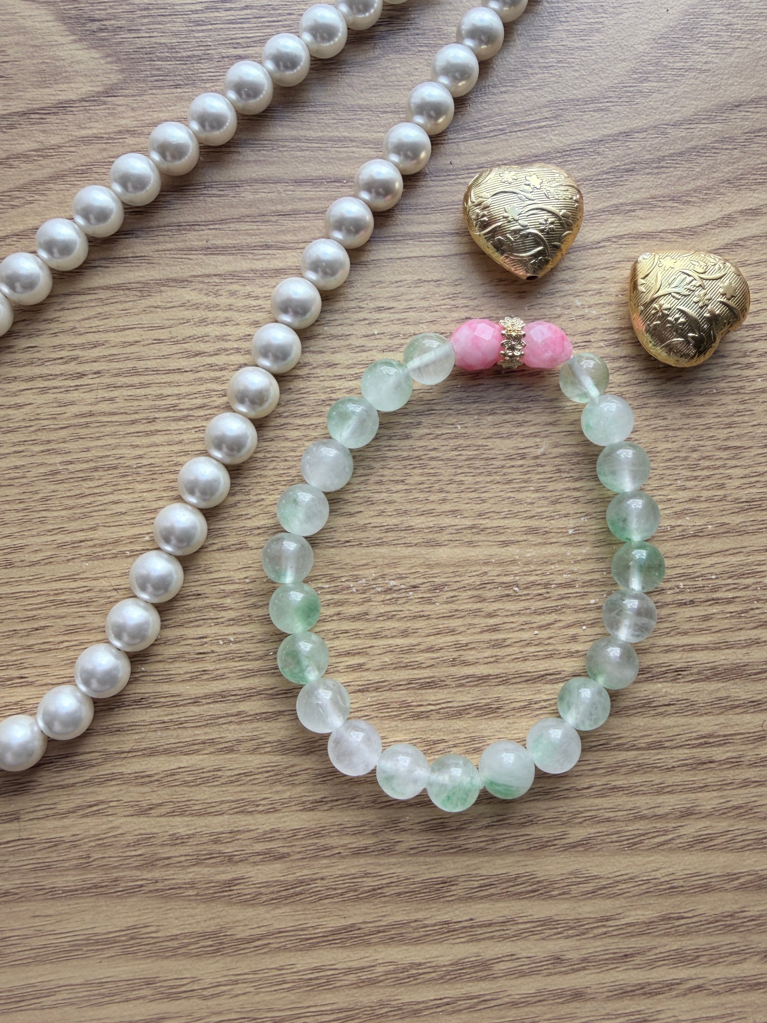 "Lifestyle shot of mint green jade and candy bracelet styled with pearl necklace and gold heart accessories on wood surface"