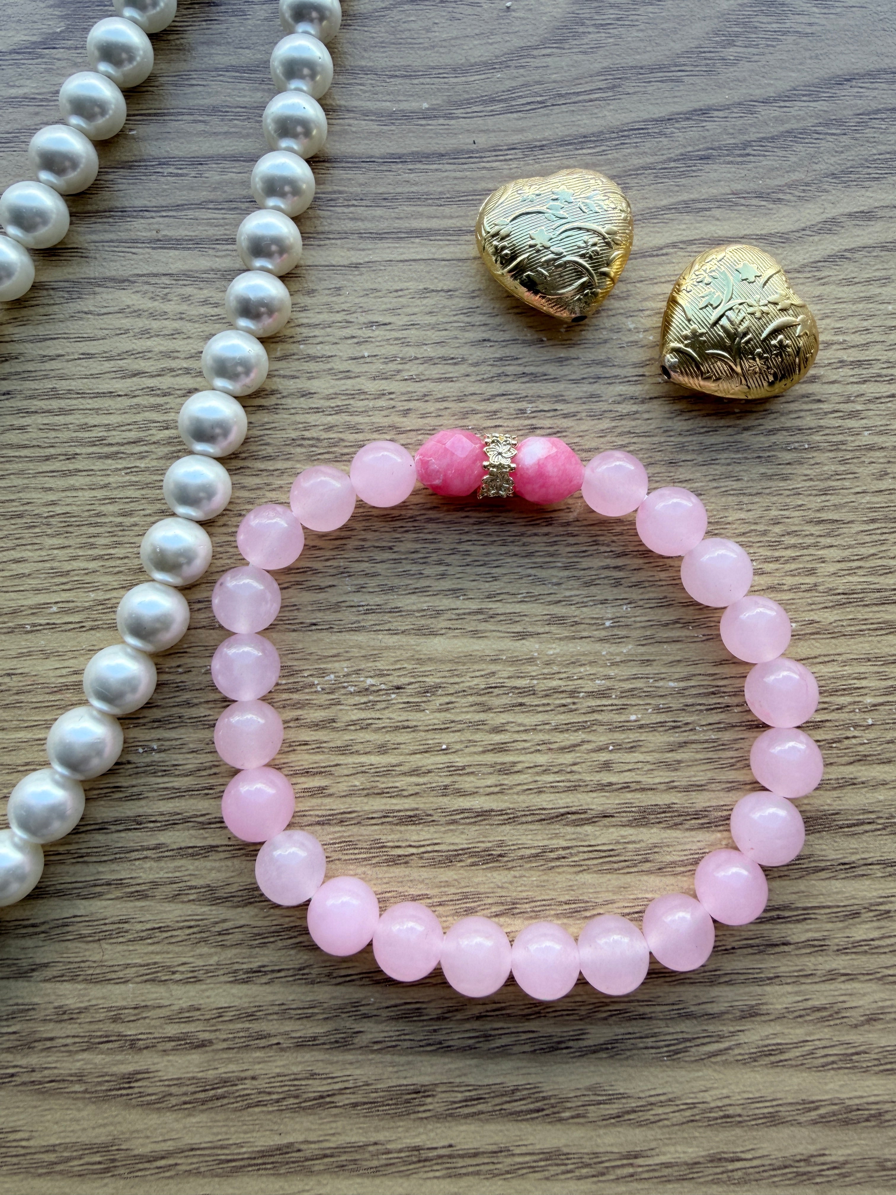 "Lifestyle shot of pink jade and candy bracelet styled with pearl necklace and gold heart accessories on wood surface"