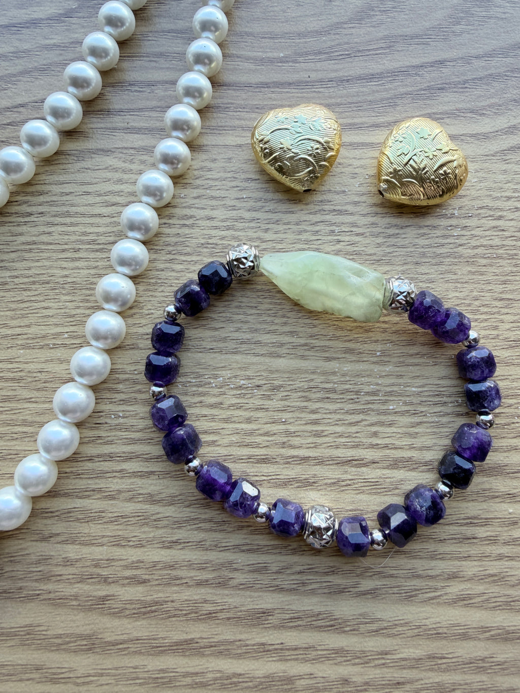 "Lifestyle shot of amethyst and green prehnite bracelet styled with pearl necklace and gold heart accessories on wood surface"