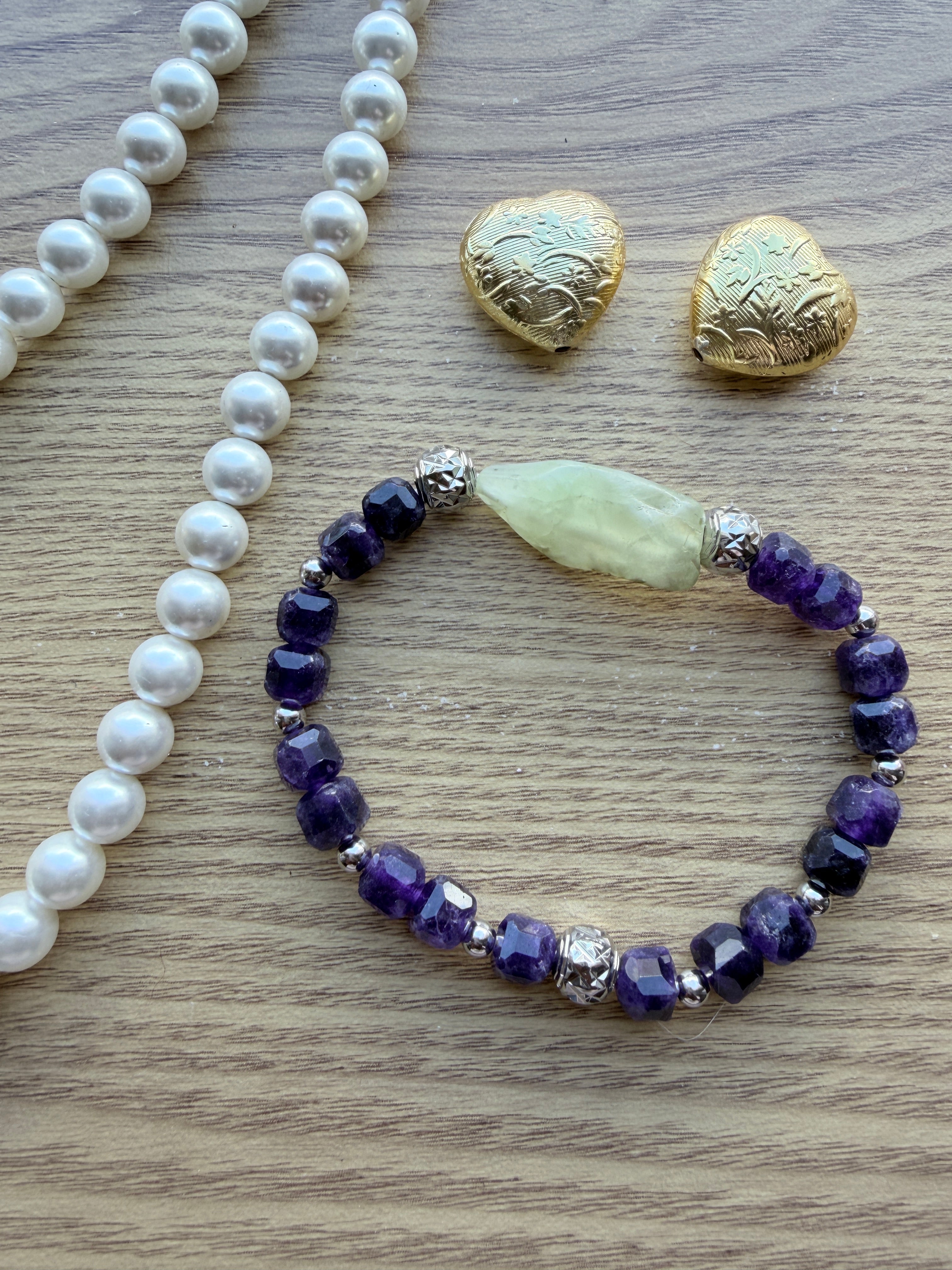 "Lifestyle shot of amethyst and green prehnite bracelet styled with pearl necklace and gold heart accessories on wood surface"