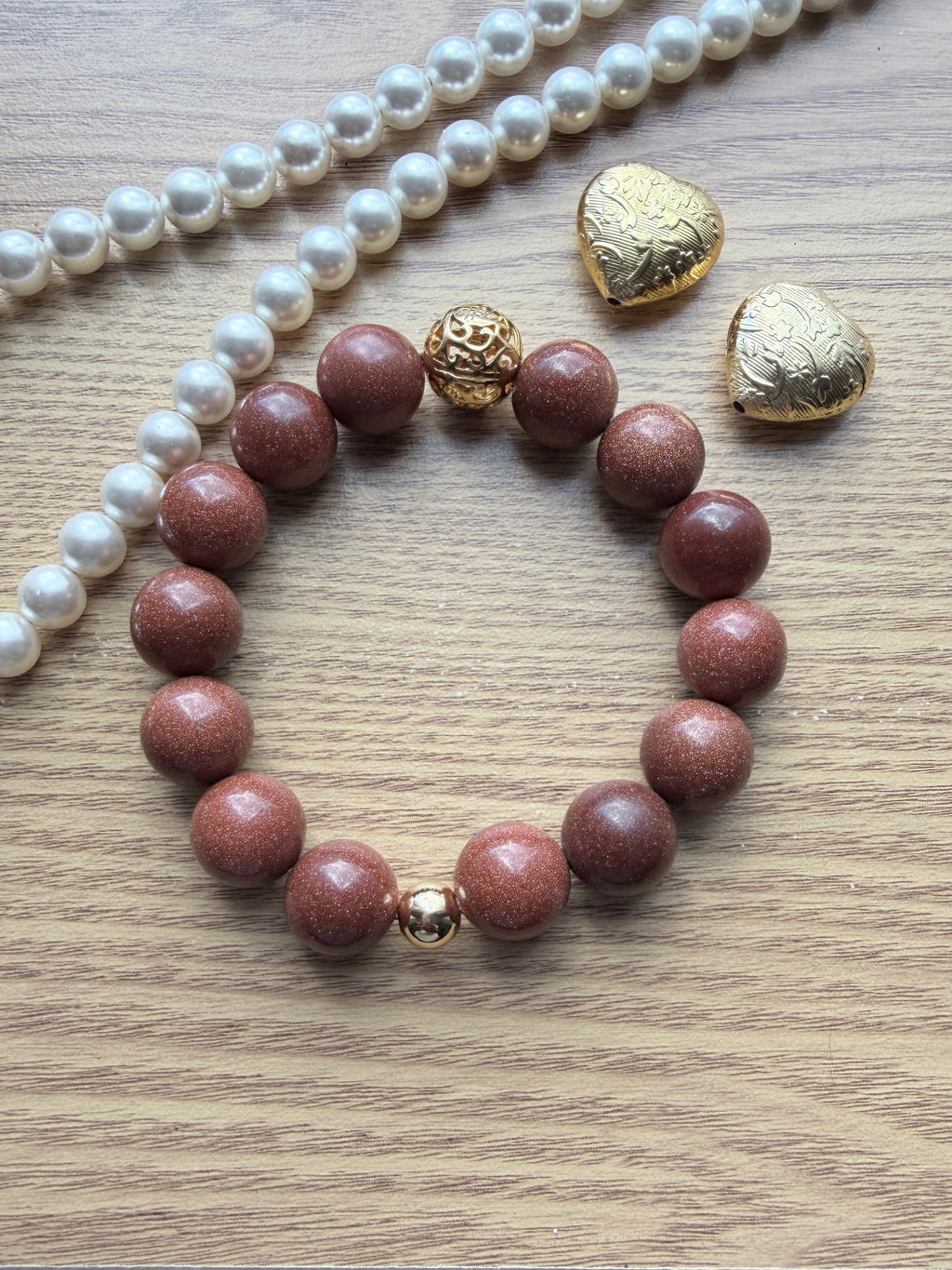 "Lifestyle shot of brown sandstone bracelet styled with pearl necklace and gold heart accessories on wood surface"