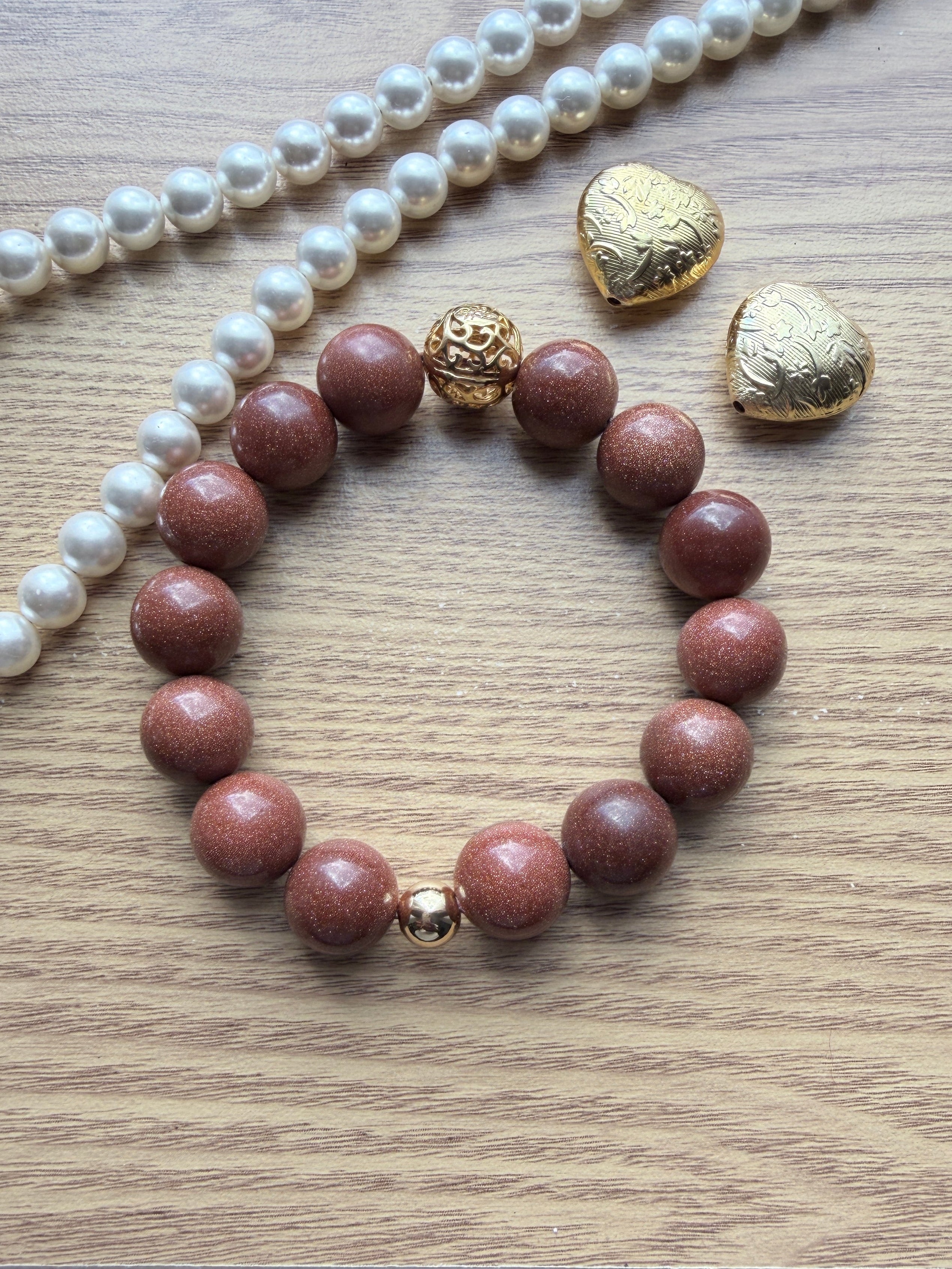 "Lifestyle shot of brown sandstone bracelet styled with pearl necklace and gold heart accessories on wood surface"