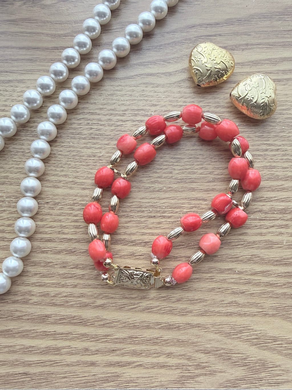 "Lifestyle shot of barrel coral two-row bracelet styled with pearl necklace and gold heart accessories on wood surface"
