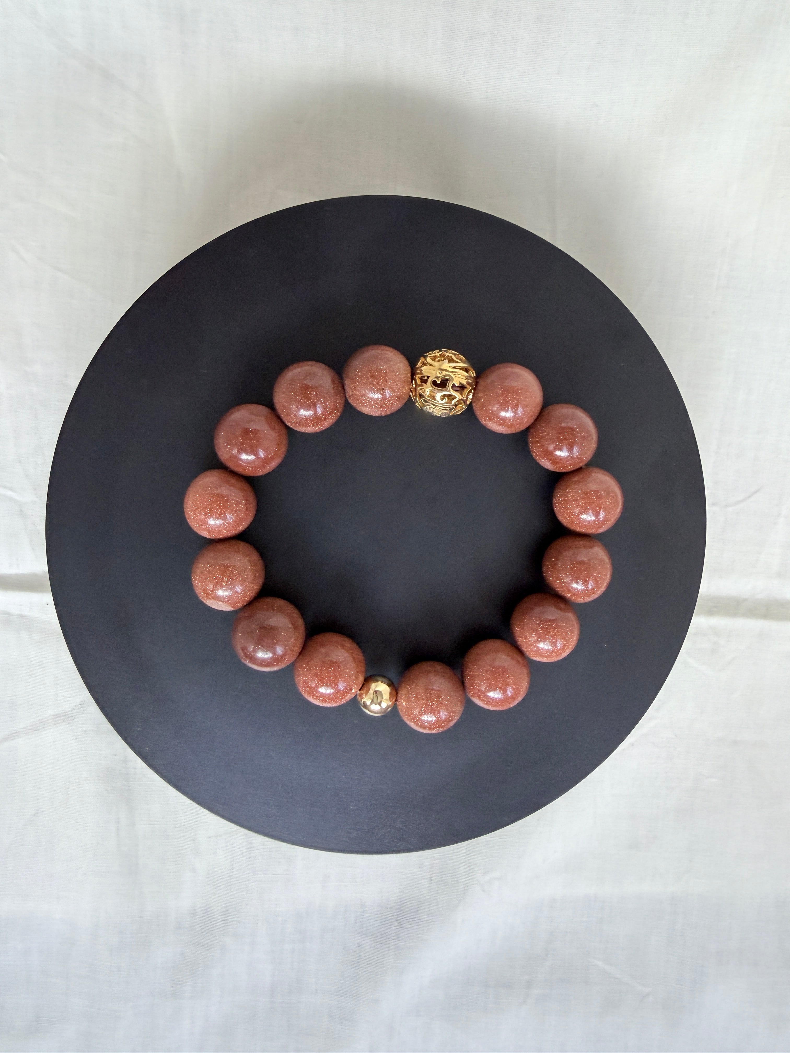 "Brown sandstone bracelet laid flat on a black circle board showing full bracelet with 14mm sparkly brown sandstone beads and ornate gold tone filigree focal bead"
