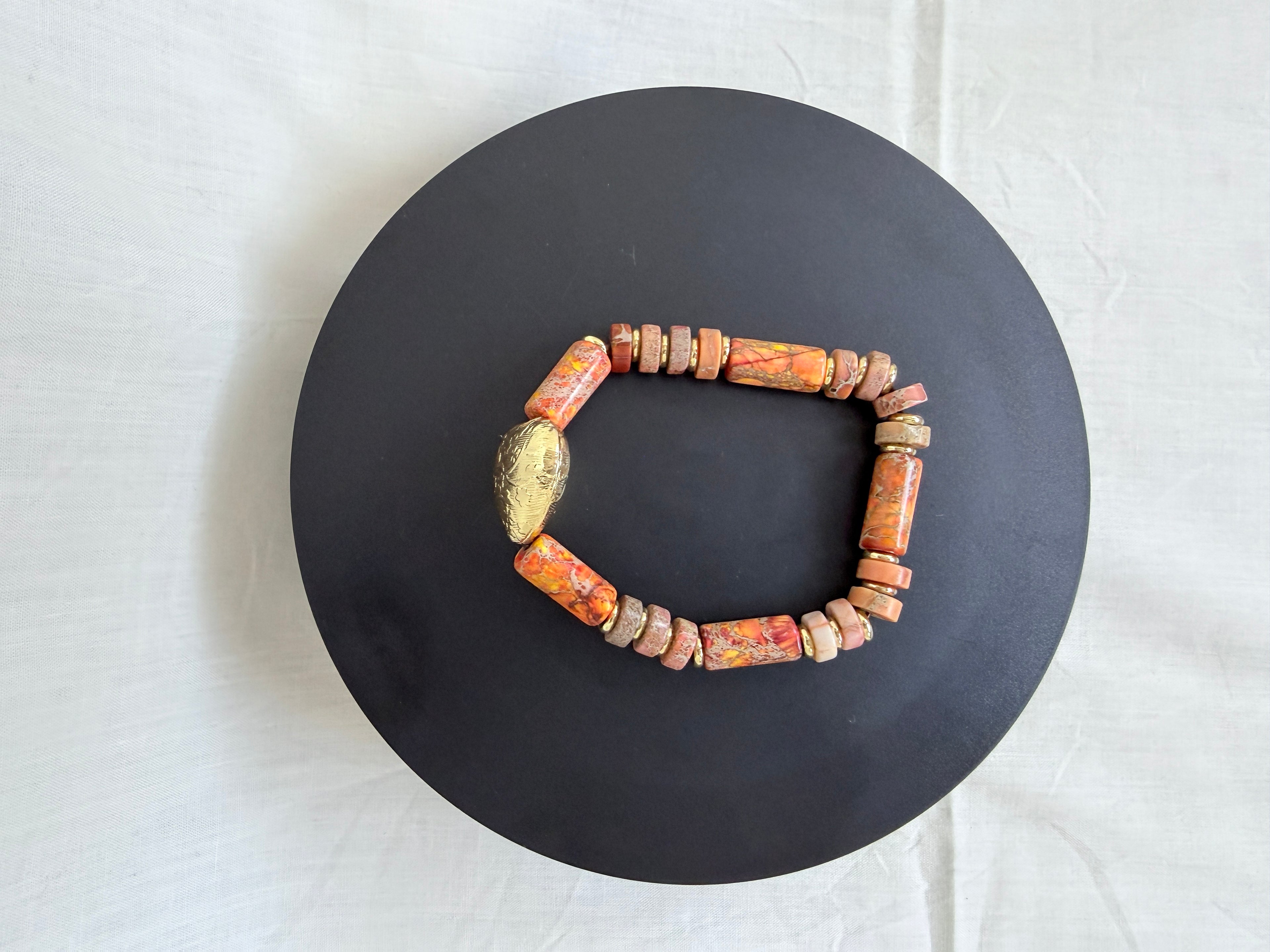 "Jasper heart bracelet laid flat on a black circle board showing full bracelet with multi-colour orange terracotta jasper tube and wheel beads and engraved brass heart focal bead"
