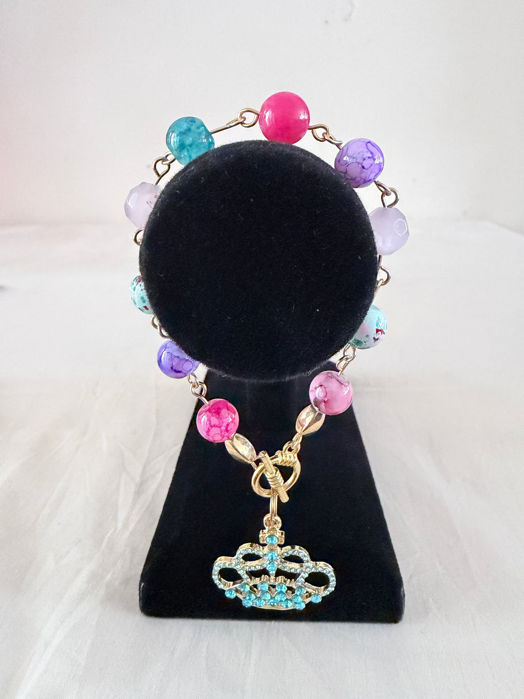 Queen of Everything Charm Bracelet on black velvet stand showing teal rhinestone gold crown charm with multicolour pink purple lavender and teal beads on gold link chain