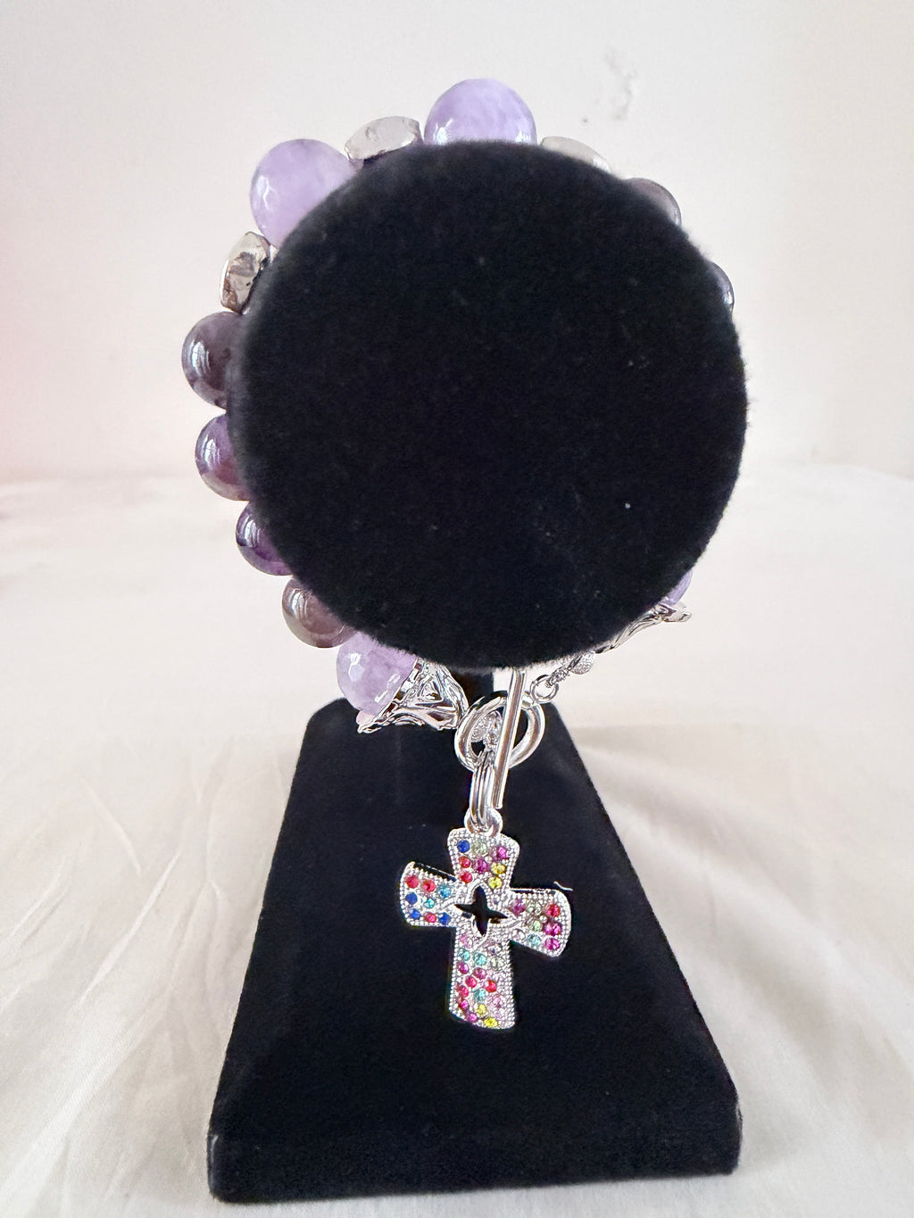Holy Grace Charm Bracelet on black velvet stand showing multicolour rhinestone silver cross charm with natural amethyst beads and silver bell end caps on toggle clasp