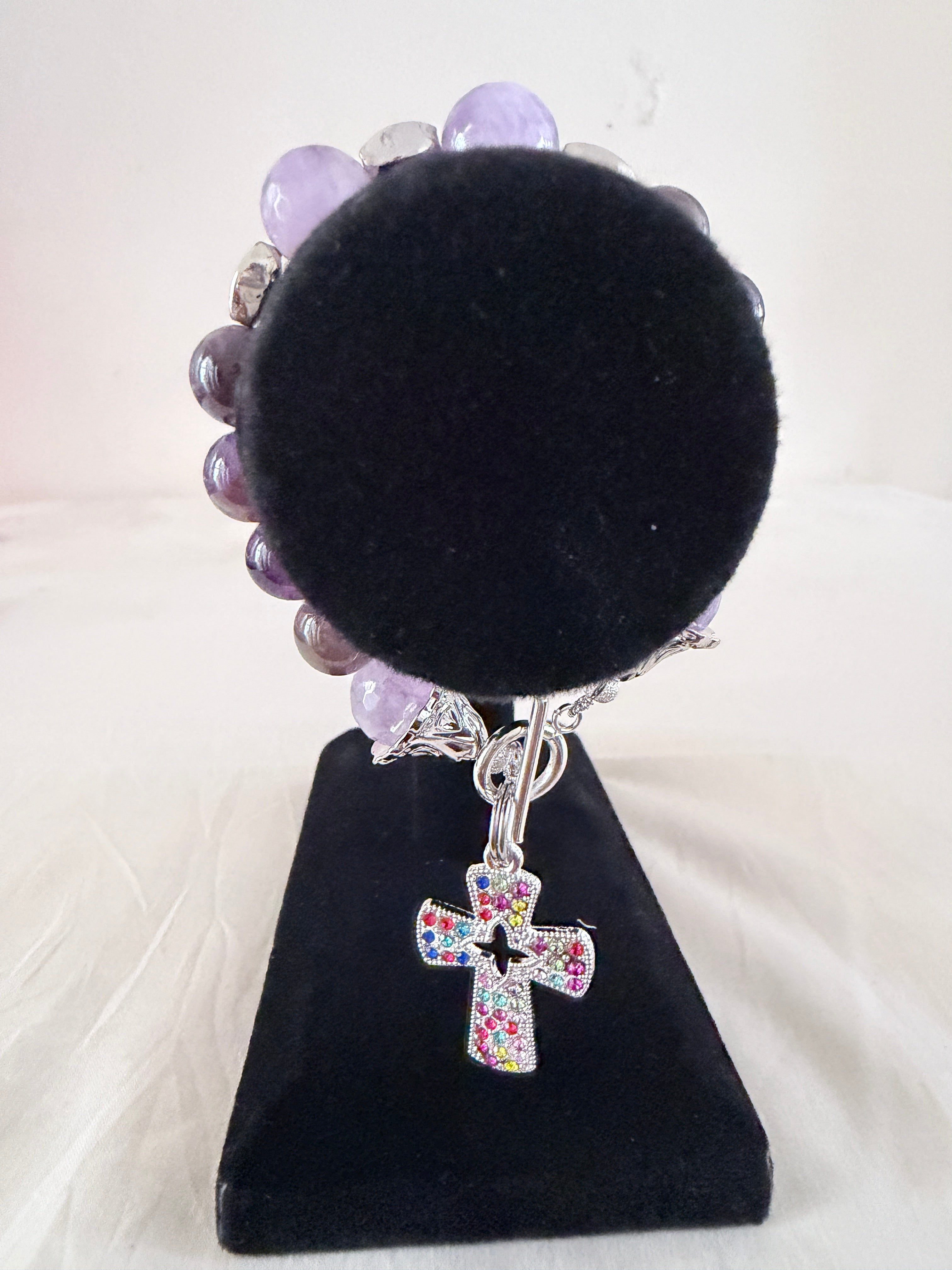 Holy Grace Charm Bracelet on black velvet stand showing multicolour rhinestone silver cross charm with natural amethyst beads and silver bell end caps on toggle clasp