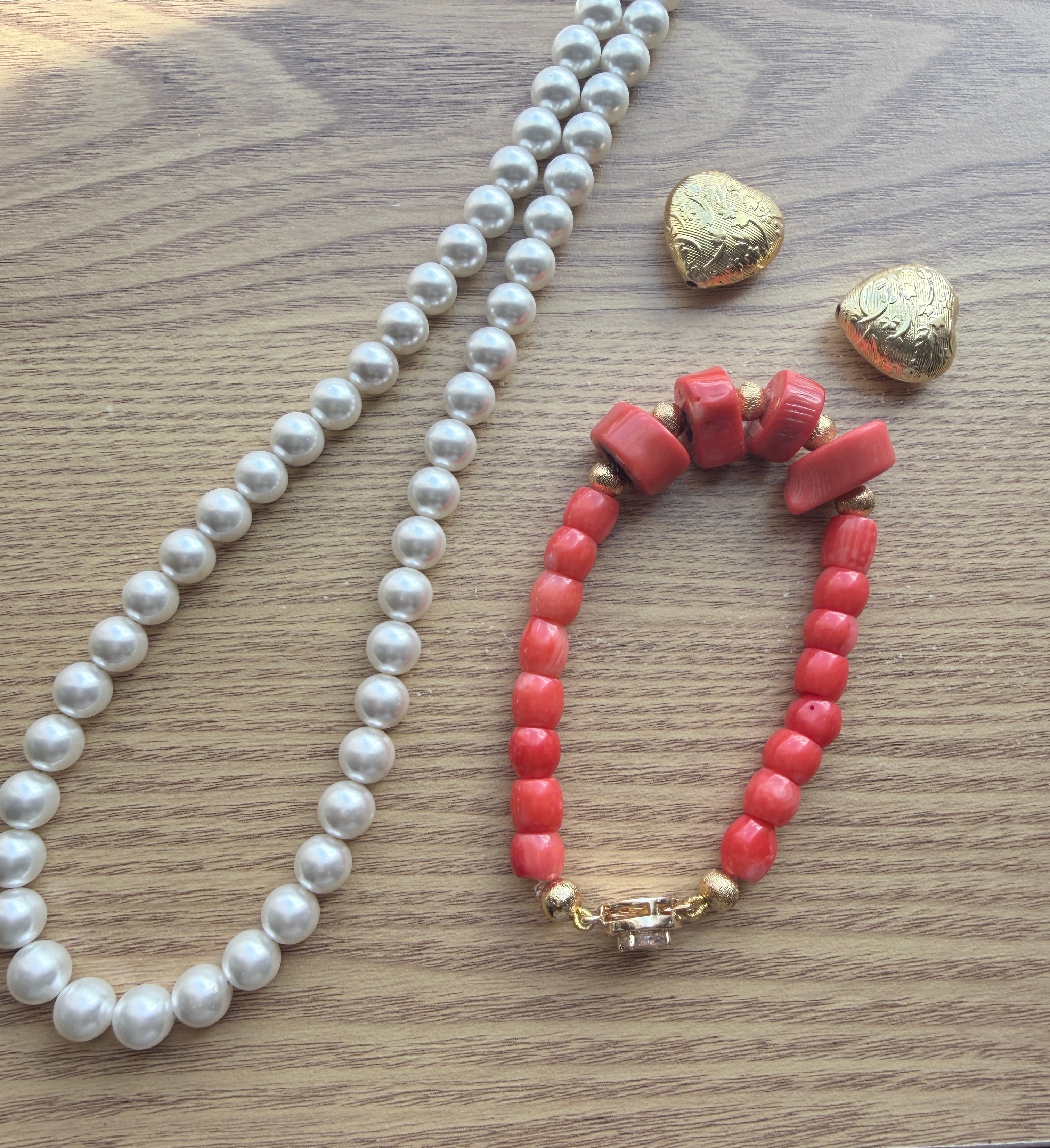 "Lifestyle shot of coral disc and barrel bracelet with CZ clasp styled with pearl necklace and gold heart accessories on wood surface"